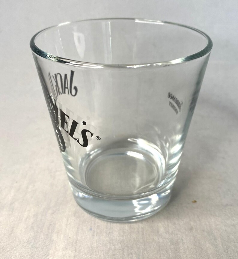 May include: A clear shot glass with the text "DANIEL'S" in black. The glass has a slightly tapered design and a thick base. The shot glass is empty and appears to be made of glass.