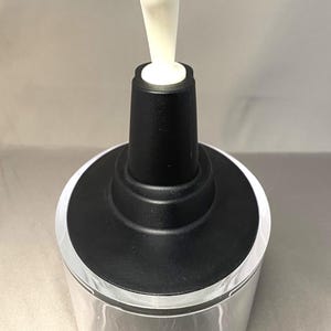 May include: A clear glass container with a black top and a white, elongated stopper. The stopper has a rounded shape. The container is cylindrical and appears to be empty. The background is a solid, light grey colour.