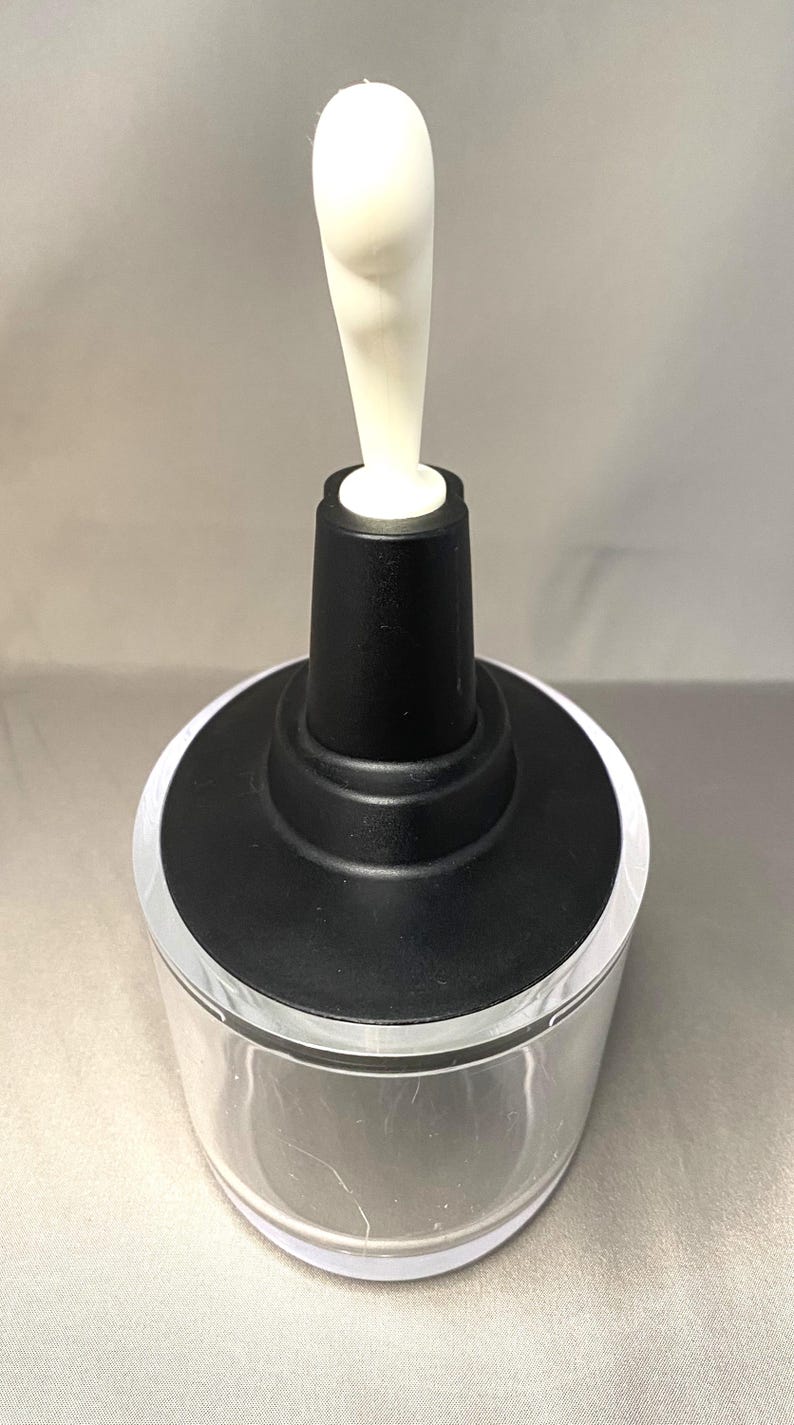 May include: A clear glass container with a black top and a white, elongated stopper. The stopper has a rounded shape. The container is cylindrical and appears to be empty. The background is a solid, light grey colour.