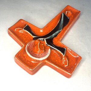 May include: A ceramic cross with a stylized figure of a person in black and white on an orange background.