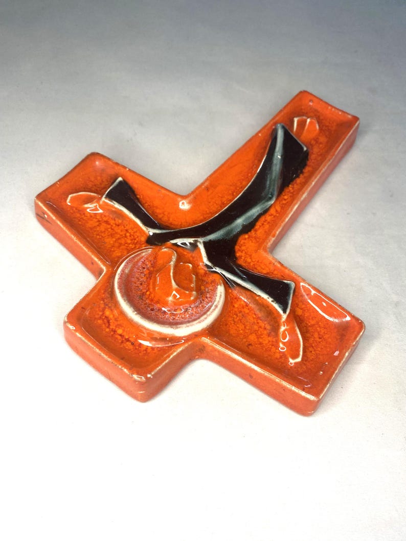 May include: A ceramic cross with a stylized figure of a person in black and white on an orange background.