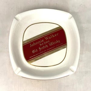 Op de afbeelding: Witte keramische asbak met een vierkante vorm en afgeronde hoeken. Het midden heeft een rood en goud label met de tekst "Johnnie Walker Red Label Old Scotch Whisky". De asbak heeft vier sigarettensteunen.