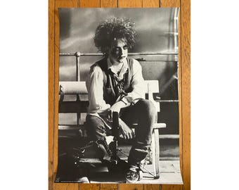 May include: Black and white photo of a person with dark hair sitting on a bench. The person is wearing a white shirt, a vest, and dark pants. The person is looking down.