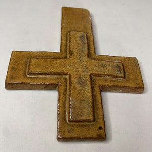 May include: A brown ceramic cross with a raised design. The cross is made of a textured clay and has a rustic look.