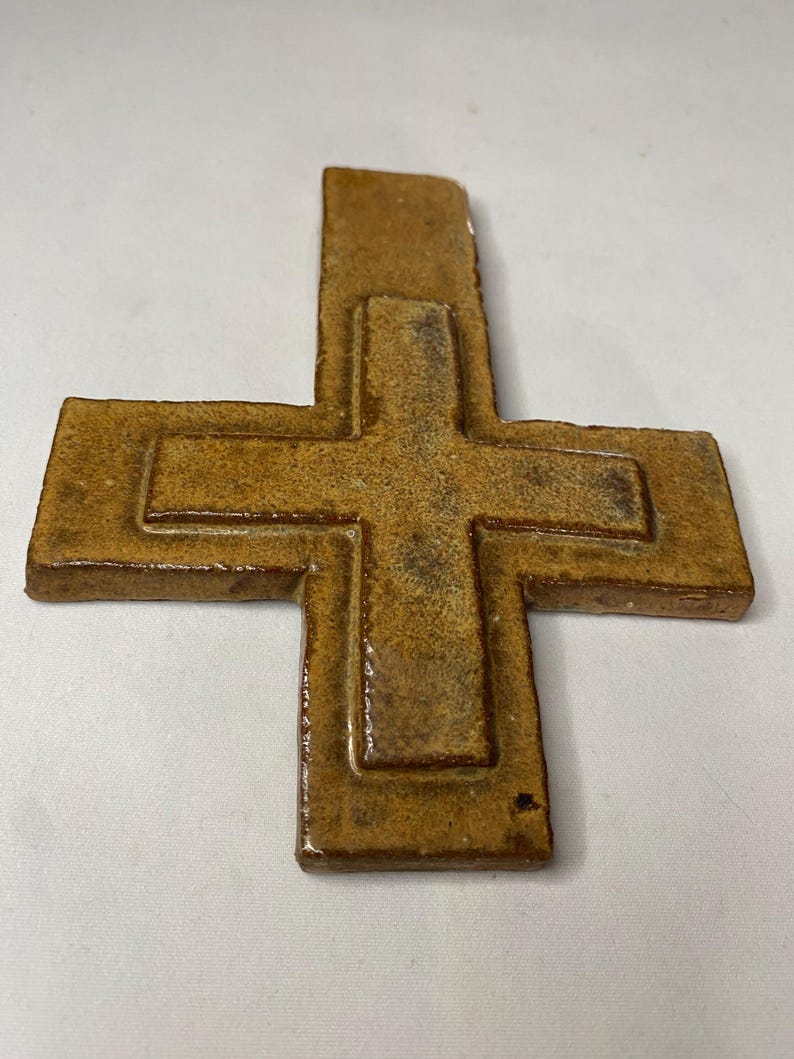 May include: A brown ceramic cross with a raised design. The cross is made of a textured clay and has a rustic look.