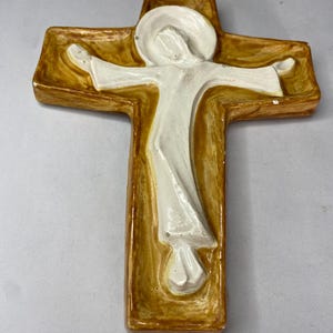 May include: A ceramic cross with a figure in white robes. The cross has a golden-brown border and a white figure with outstretched arms. The figure has a halo. The cross is a religious item.