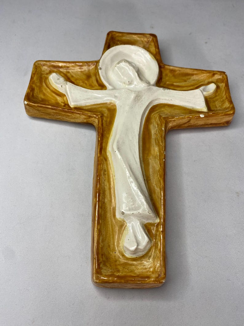 May include: A ceramic cross with a figure in white robes. The cross has a golden-brown border and a white figure with outstretched arms. The figure has a halo. The cross is a religious item.