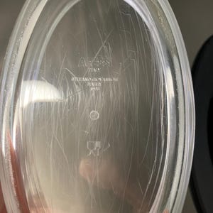 May include: Clear, oval-shaped dish with visible scratches. The dish has the text "Alessi ITALY" and other text that is difficult to read. The dish is made of a transparent material.