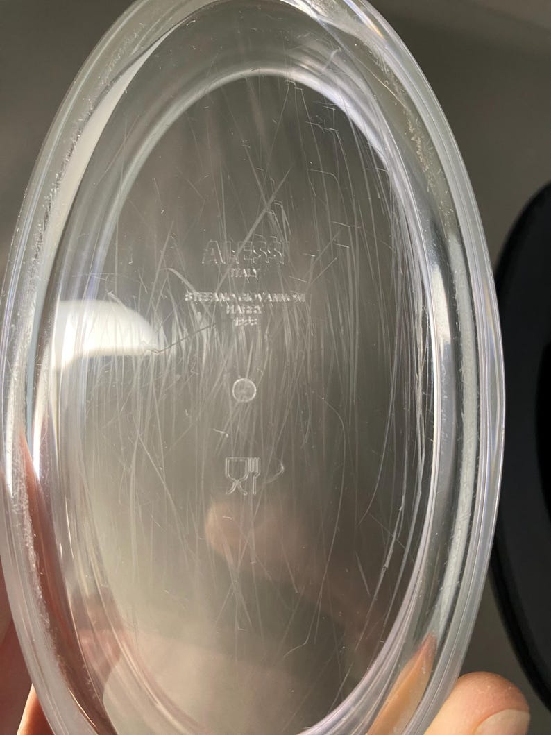 May include: Clear, oval-shaped dish with visible scratches. The dish has the text "Alessi ITALY" and other text that is difficult to read. The dish is made of a transparent material.
