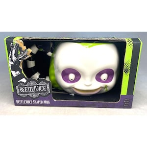 Könnte beinhalten: Eine weiß-grün-lila Beetlejuice-Tasse in Originalverpackung. Die Tasse zeigt ein Cartoon-Gesicht mit lila Augen und einem grünen Fleck. Auf der Box sind das Beetlejuice-Logo und der Schriftzug "Beetlejuice Shaped Mug" zu sehen.