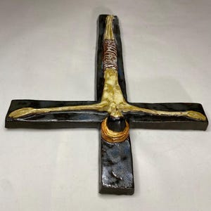 Może przedstawiać: Ceramiczna rzeźba ścienna przedstawiająca szkieletową postać na czarnym krzyżu. Postać jest umieszczona do g&oacute;ry nogami, z głową na dole krzyża. Krzyż jest ozdobiony niebieską i brązową glazurą.