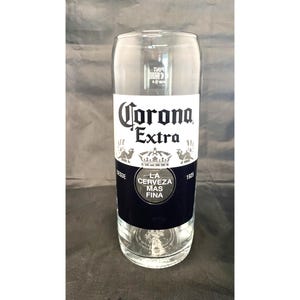 Könnte beinhalten: Ein klares Bierglas mit dem "Corona Extra"-Logo in Weiß auf einem schwarz-weißen Etikett. Das Glas hat eine zylindrische Form und ist für Bier konzipiert.