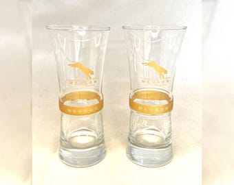 May include: Two clear glass shot glasses with a gold band around the middle. The glasses have a gold panther logo and the text "MEUKOW COGNAC" printed on them.
