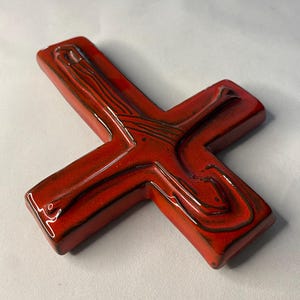 May include: A glossy, red ceramic cross with a textured surface and dark brown accents. The cross features a stylized design with curved lines and a smooth, reflective finish. The cross is a religious symbol.