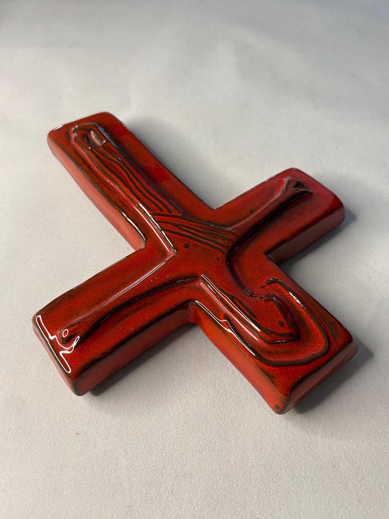 May include: A glossy, red ceramic cross with a textured surface and dark brown accents. The cross features a stylized design with curved lines and a smooth, reflective finish. The cross is a religious symbol.