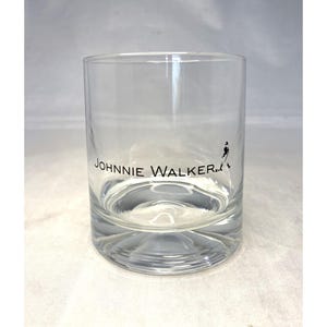 May include: A clear, cylindrical rocks glass with the "JOHNNIE WALKER" logo in black. The logo includes the brand name and a silhouette of the Striding Man. The glass is designed for serving whiskey or other spirits.