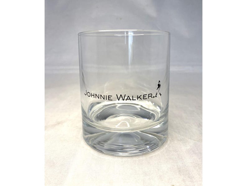 May include: A clear, cylindrical rocks glass with the "JOHNNIE WALKER" logo in black. The logo includes the brand name and a silhouette of the Striding Man. The glass is designed for serving whiskey or other spirits.