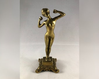 May include: A gold-toned metal figurine of a woman standing on a decorative base. The woman is depicted with her arms raised, as if she is flexing her muscles.