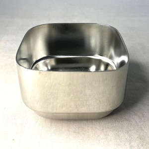 May include: A small, square, silver-coloured container with rounded corners. The container has a smooth, reflective surface and a slightly curved base. The interior is also silver and reflects the light. The container is empty.