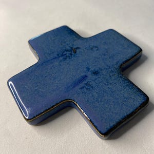 K&ouml;nnte beinhalten: Ein blaues Keramikkreuz mit dunkelbrauner Glasur. Das Kreuz ist leicht erh&ouml;ht und hat eine abgerundete Form.