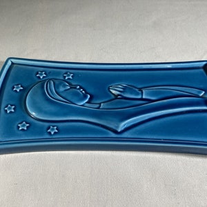 May include: A blue ceramic wall plaque with a raised image of a woman lying down with her hands raised. The plaque is decorated with stars.