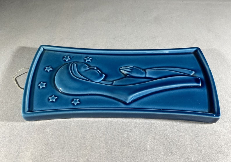 May include: A blue ceramic wall plaque with a raised image of a woman lying down with her hands raised. The plaque is decorated with stars.
