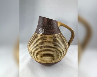May include: A brown and beige ceramic pitcher with a geometric pattern. The pitcher has a handle and a small silver label with a symbol on it.