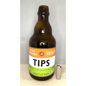 Könnte beinhalten: Eine braune Bierflasche mit einem weißen, grünen und orangefarbenen Etikett. Auf dem Etikett steht "EXTRA ORDINARY VEDETT TIPS" mit zusätzlichem Text. Ein kleines, graues Feuerzeug befindet sich neben der Flasche.