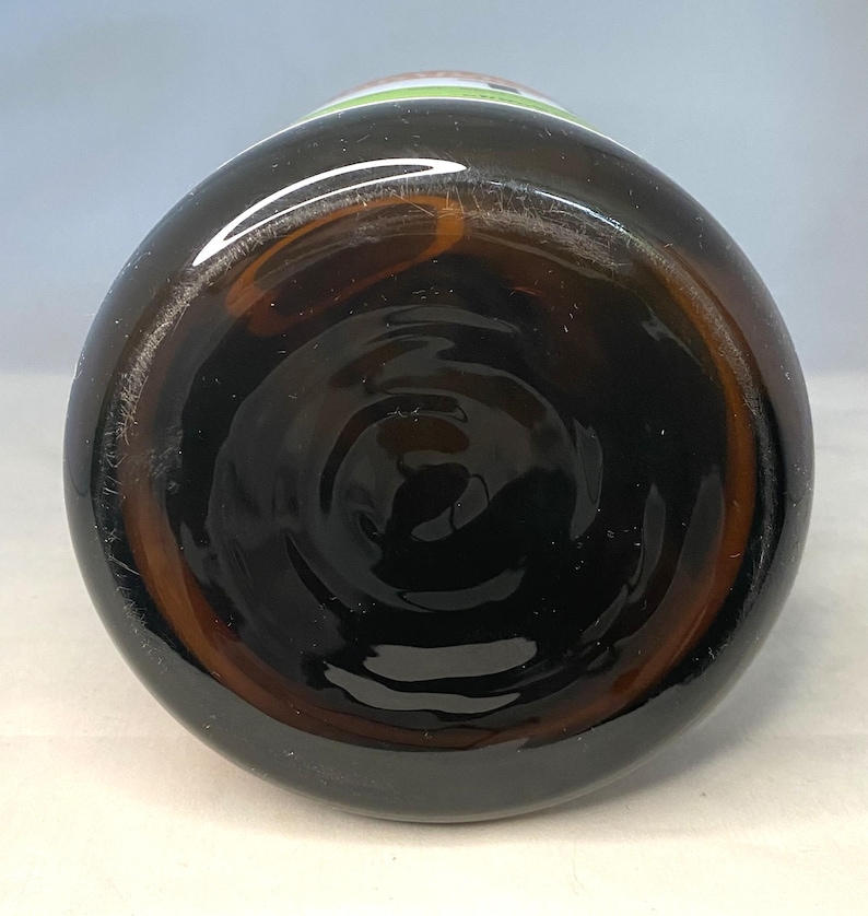May include: A brown glass bottle, viewed from the bottom, showcasing a dark, swirling interior. The bottle's base has a circular design, and the glass has a slight amber hue. The top of the bottle has a green and white label.