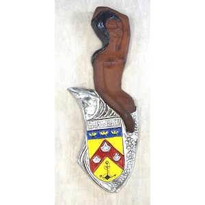 May include: A silver and brown mermaid figurine with a crest that says "topke-heist" and features a yellow anchor and three red scallop shells.