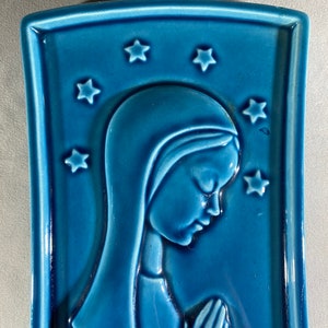 May include: A blue ceramic wall plaque with a raised image of a woman in prayer. The woman is wearing a blue robe and has her hands clasped in front of her. There are stars around the woman's head.