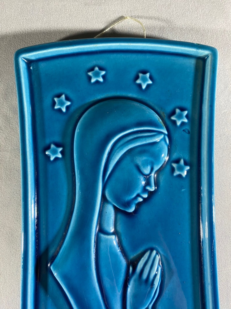 May include: A blue ceramic wall plaque with a raised image of a woman in prayer. The woman is wearing a blue robe and has her hands clasped in front of her. There are stars around the woman's head.