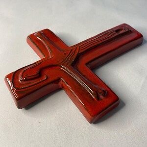 May include: A red ceramic cross with a glossy finish. The cross features a detailed depiction of a figure, with brown accents. The religious symbol is a decorative item.