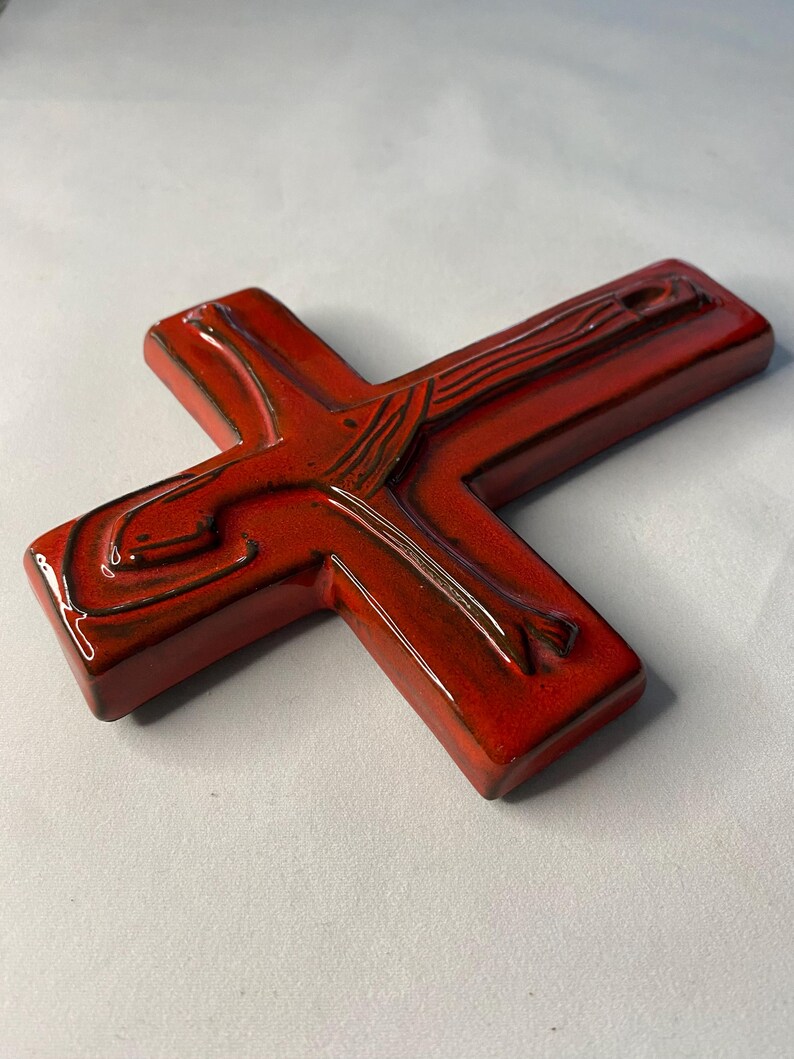May include: A red ceramic cross with a glossy finish. The cross features a detailed depiction of a figure, with brown accents. The religious symbol is a decorative item.