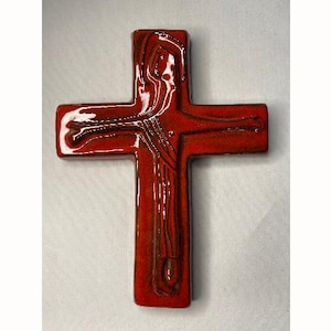 May include: A glossy, red ceramic cross with a detailed depiction of a figure. The figure is outlined in dark brown, with the cross itself having a rectangular shape. The cross is a religious symbol.