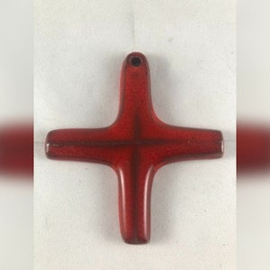 May include: A red ceramic cross with a glossy finish. The cross has a hole at the top for hanging. The design features a traditional Christian cross shape with rounded edges. The color is a deep, rich red.