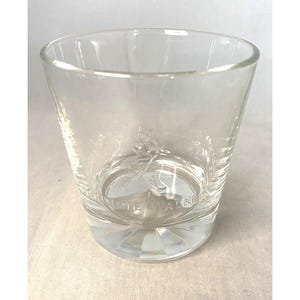 Puede incluir: Un vaso de vidrio transparente y vac&iacute;o con boca ancha y base gruesa estampada. El vaso tiene una forma ligeramente acampanada y un logotipo visible en la parte inferior. El vaso est&aacute; dise&ntilde;ado para beber.