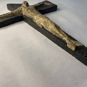 May include: A black wooden crucifix with a beige figure of a person on it. The figure is sculpted in a realistic style and has a textured surface.