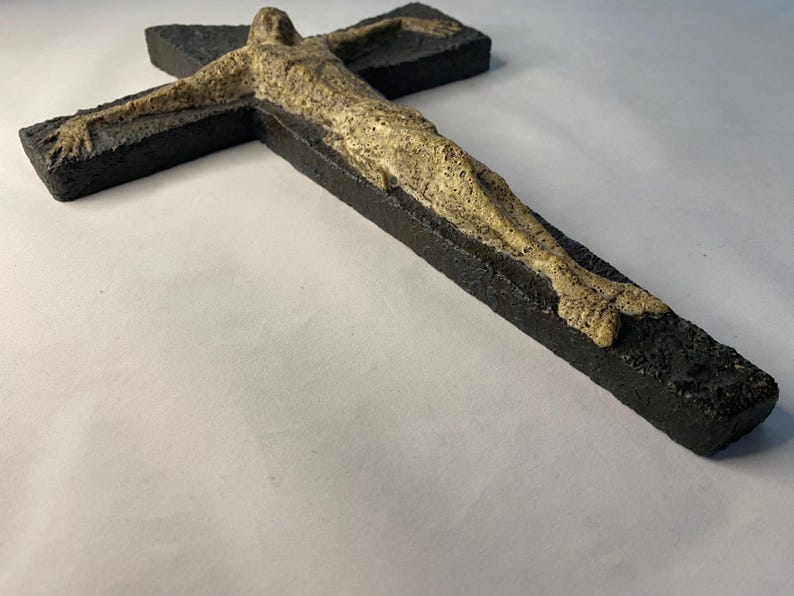 May include: A black wooden crucifix with a beige figure of a person on it. The figure is sculpted in a realistic style and has a textured surface.