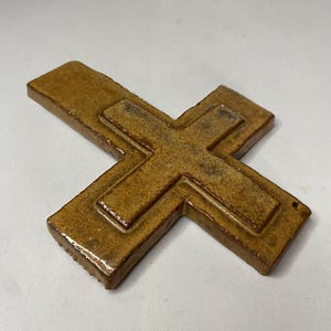 May include: A brown ceramic cross with a raised design. The cross is made of a textured clay and has a rustic look.