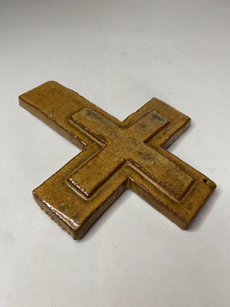 May include: A brown ceramic cross with a raised design. The cross is made of a textured clay and has a rustic look.