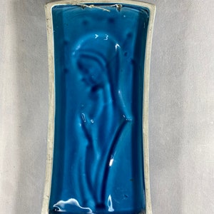 May include: A blue ceramic wall plaque with a raised design of a stylized flower. The plaque has a white border and a string for hanging.