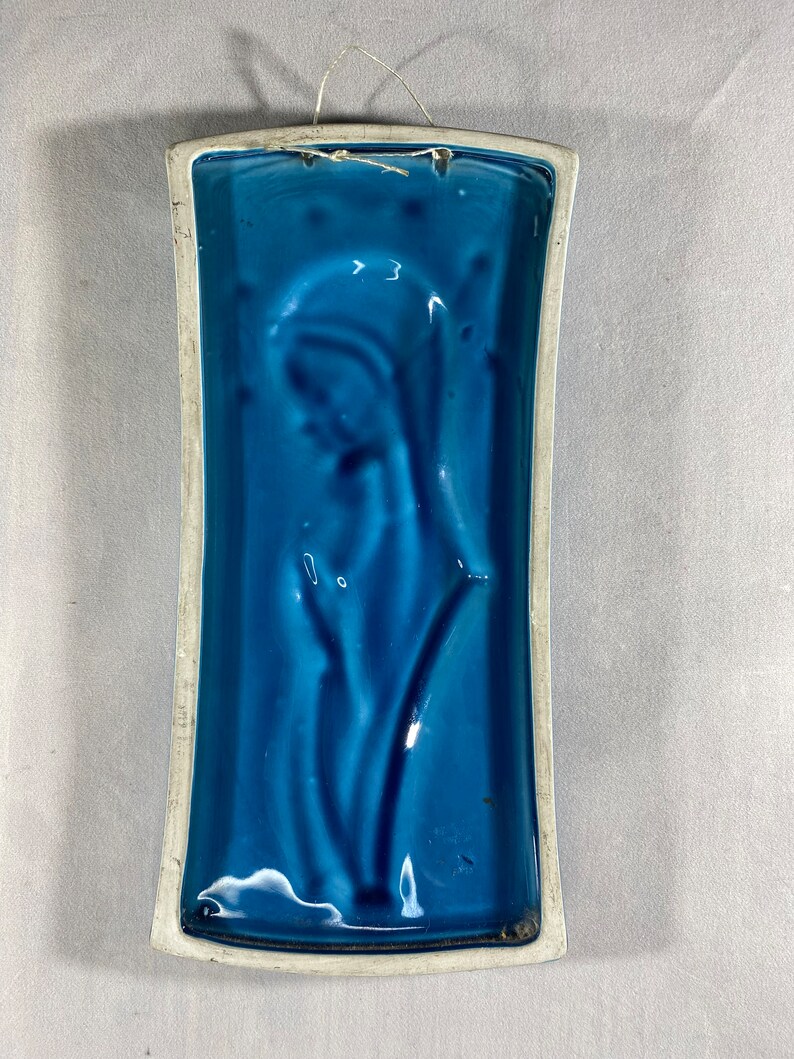 May include: A blue ceramic wall plaque with a raised design of a stylized flower. The plaque has a white border and a string for hanging.