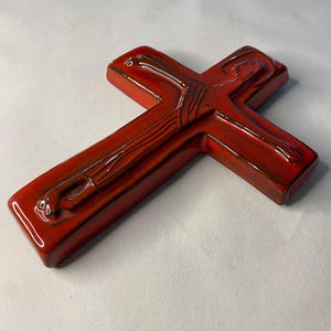 May include: A glossy, red ceramic cross with dark brown detailing. The cross has a smooth, rounded shape and a high-gloss finish. The design features subtle lines and curves, adding depth to the religious symbol.