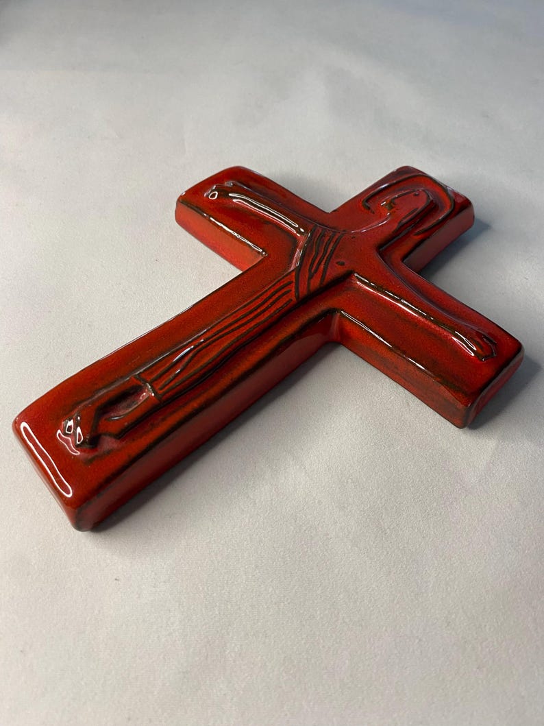 May include: A glossy, red ceramic cross with dark brown detailing. The cross has a smooth, rounded shape and a high-gloss finish. The design features subtle lines and curves, adding depth to the religious symbol.