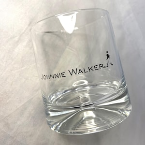 May include: A clear, lowball glass with the text "JOHNNIE WALKER" printed in black. A black silhouette of a man is walking on the right side of the glass. The glass is empty and sits on a white surface.