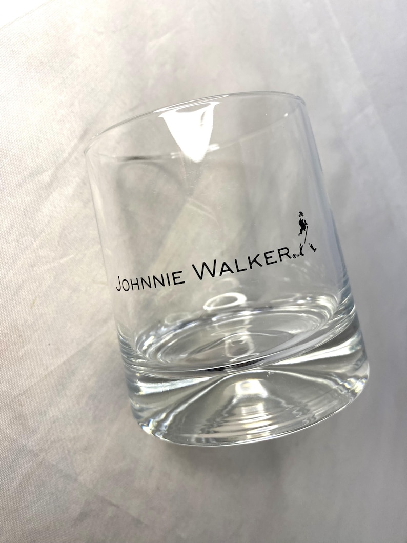 May include: A clear, lowball glass with the text "JOHNNIE WALKER" printed in black. A black silhouette of a man is walking on the right side of the glass. The glass is empty and sits on a white surface.