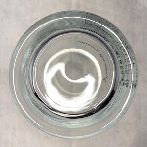 May include: Clear glass tumbler with a wide rim, viewed from above. The bottom of the glass has a slight curve. Faint text is visible around the rim. The glass is sitting on a light gray surface.