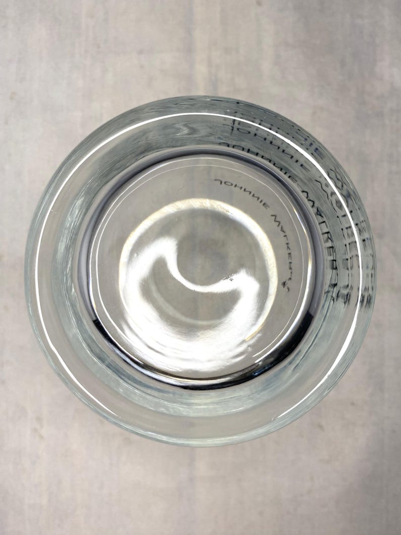 May include: Clear glass tumbler with a wide rim, viewed from above. The bottom of the glass has a slight curve. Faint text is visible around the rim. The glass is sitting on a light gray surface.