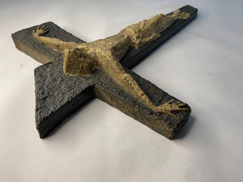 May include: A dark gray ceramic cross with a raised, light brown figure of a person on it.